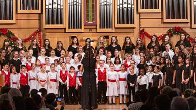 A large group of children stand on risers in front of a church organ as they sing.