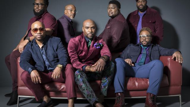 seven black men sit on a red leather couch looking at the camera