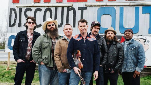 seven men standing in jeans and shirts and cowboy hats looking at the camera