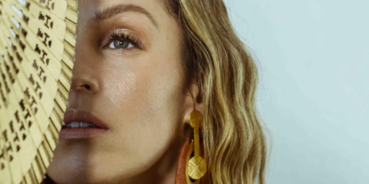 Caucasian woman wearing gold attire, brown earrings and holding a fan covering half her face.