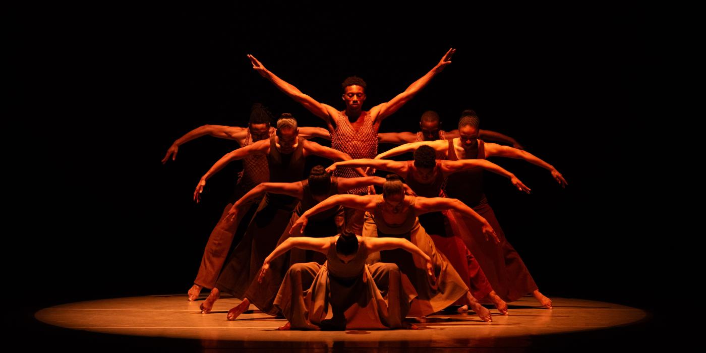 Group of 9 dancers under a spotlight with arms outstretched wide