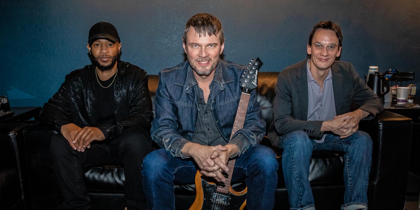Three men sit on a couch smiling at the camera. One holds a guitar.