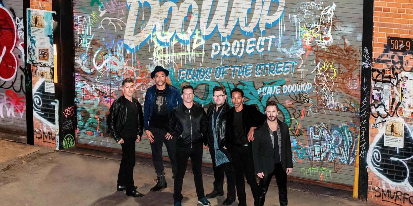 Six men standing in front  of a graffiti wall.