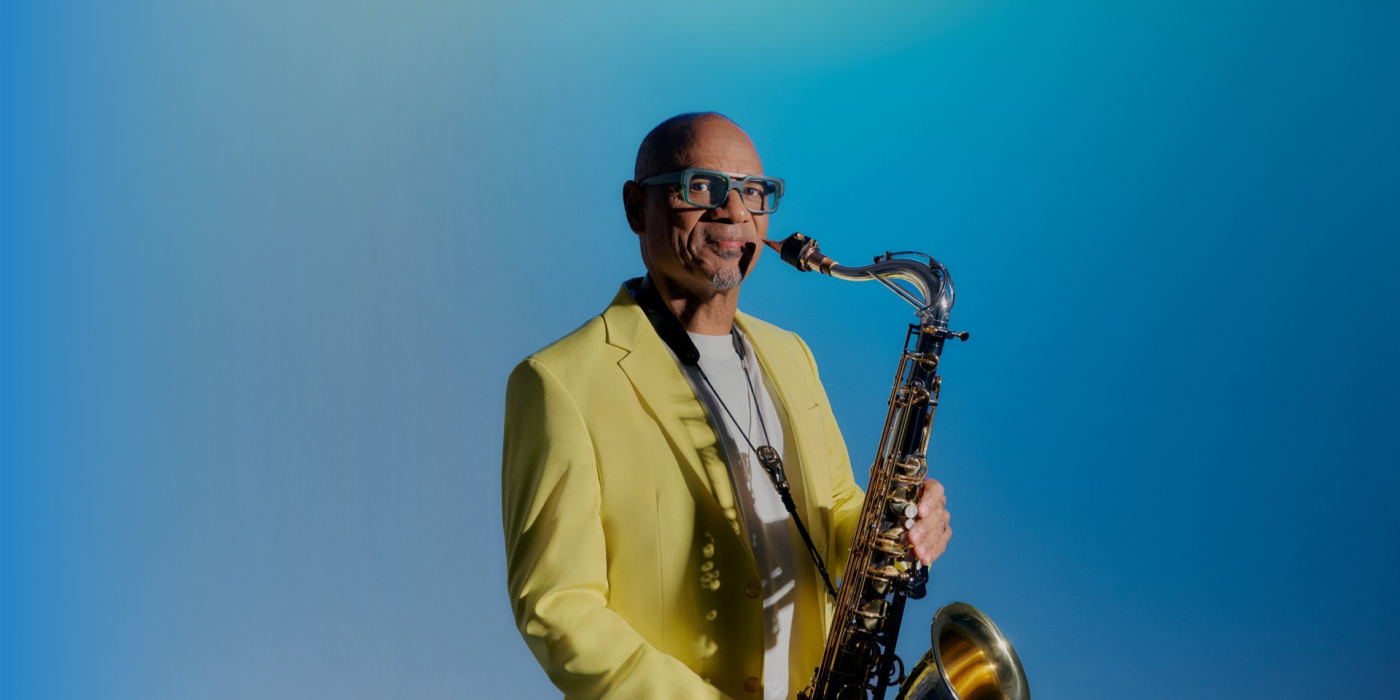 African American man wearing a yellow blazer holding a saxophone.