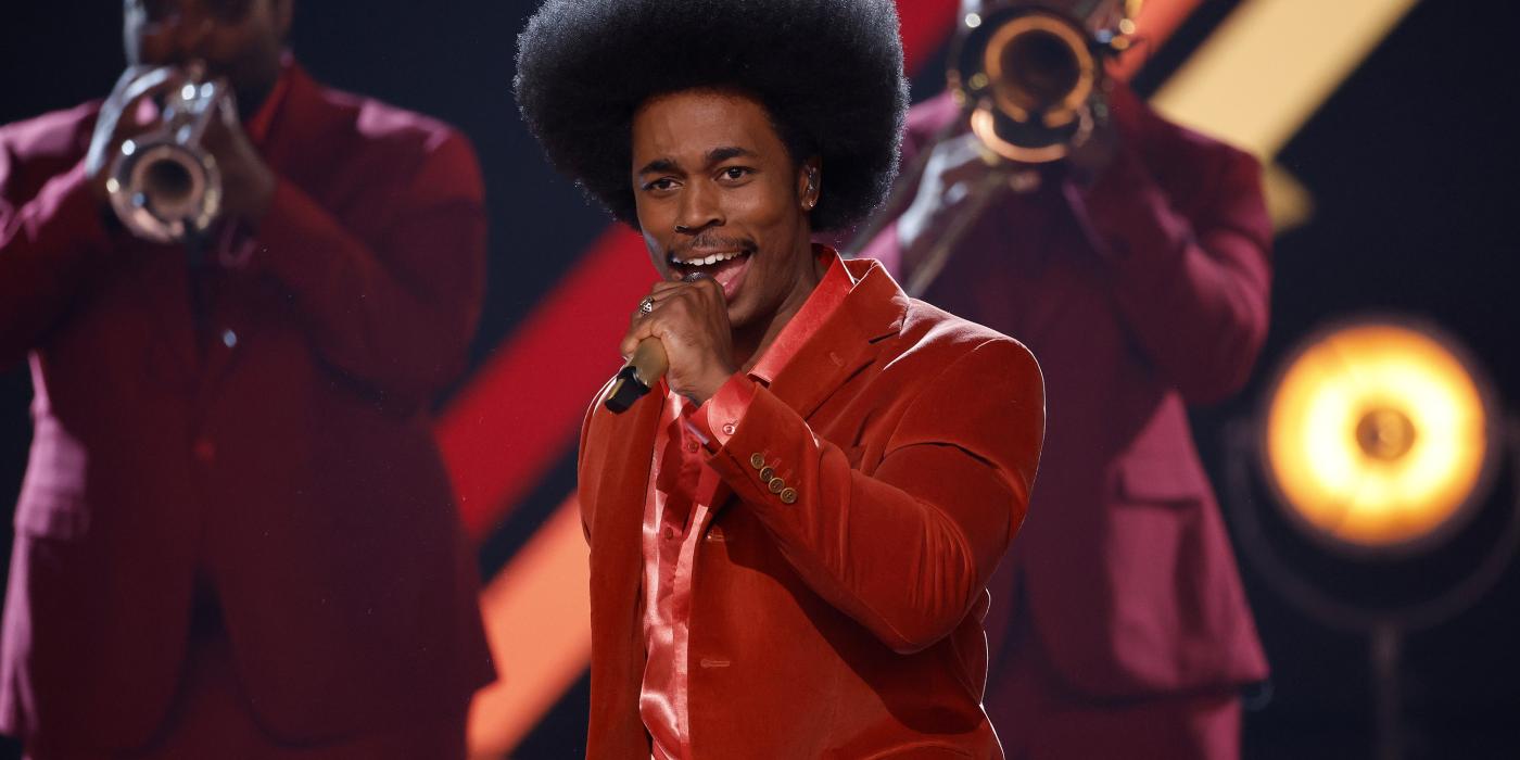 singer dressed in red singing on stage with trumpeters in background