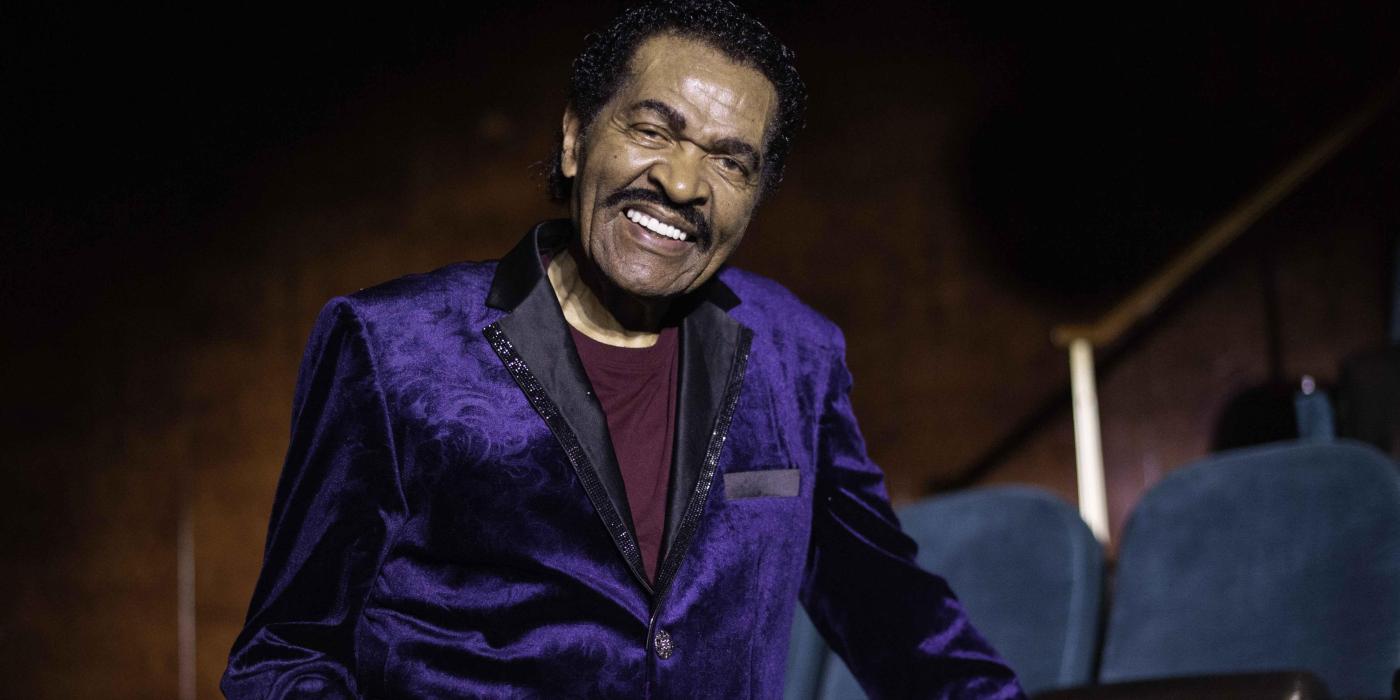 A man with short dark hair and a mustache, smiles at the camera. He wears a purple sport coat