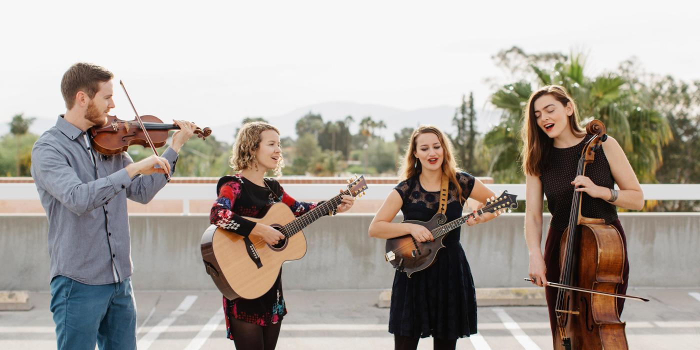 Three women and a man, sing and play their string instruments on the roof of a parking garage on a sunny day