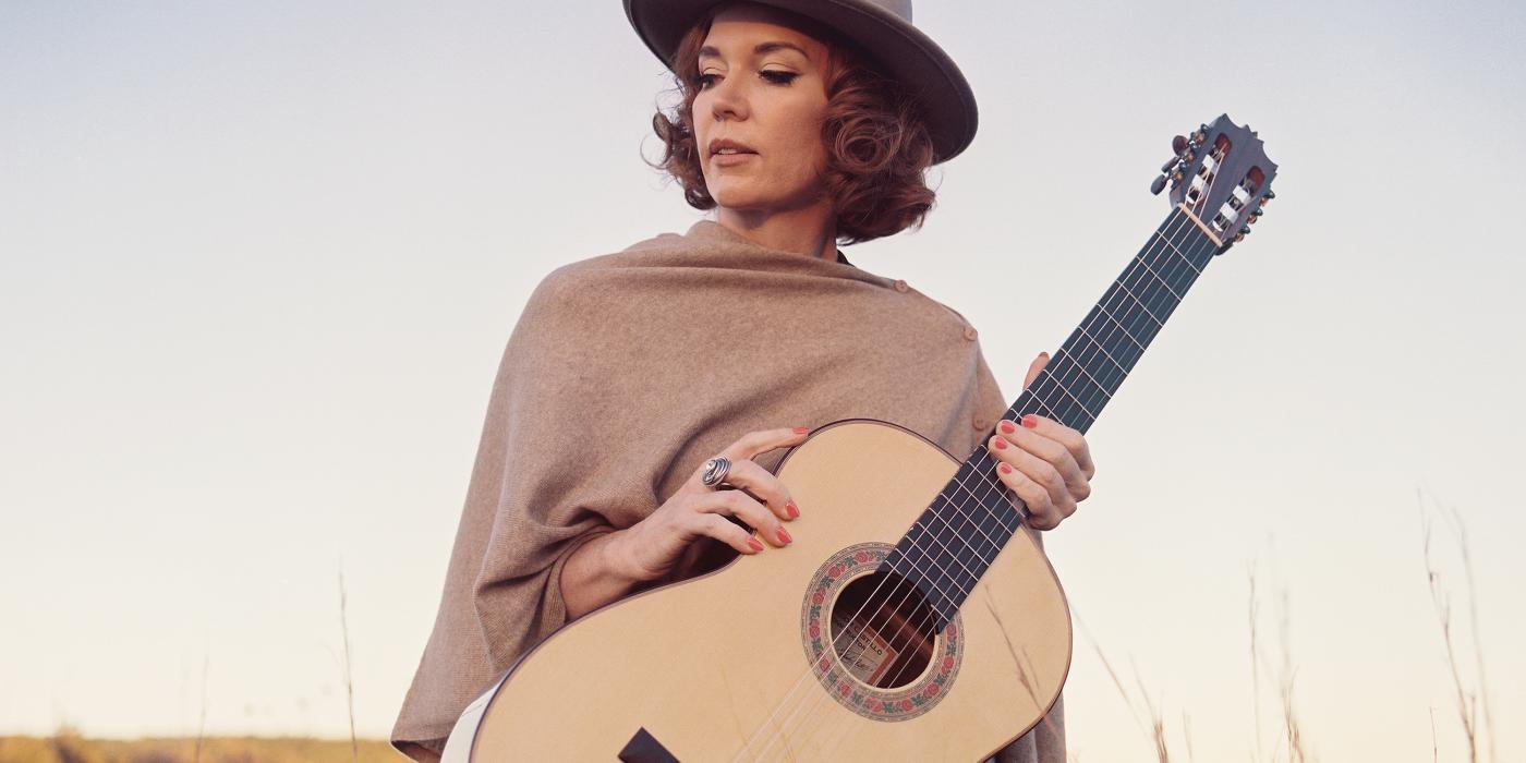 Sue Foley in field of grass looking off to the side in a hat and shall holding an acoustic guitar