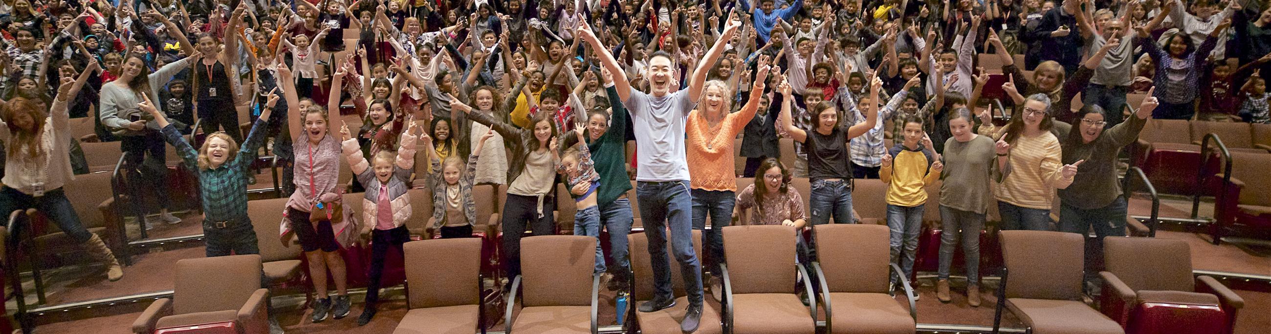 Artist Alpin Hong stands with his hands raised in joy along with a theatre full of children
