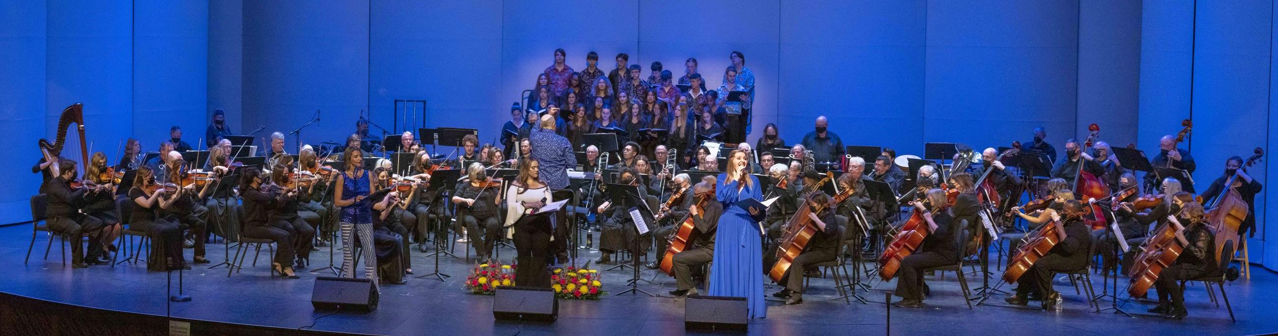 A full orchestra of musicians, conductor and two singers perform on the stage,
