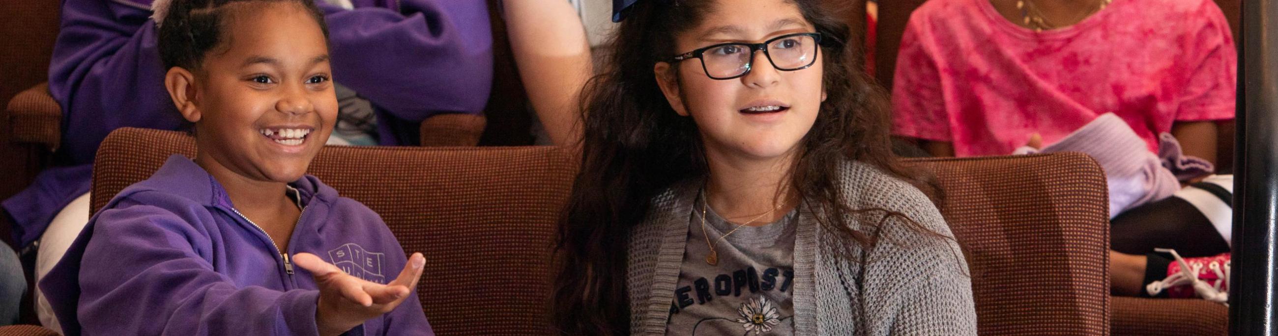 Two young girls sit in theatre seats, enjoying a show