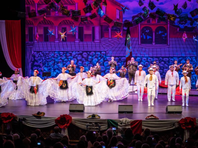 Folklorico dancers dressed in white costumes dance on a stage with mariachi musicians behind them