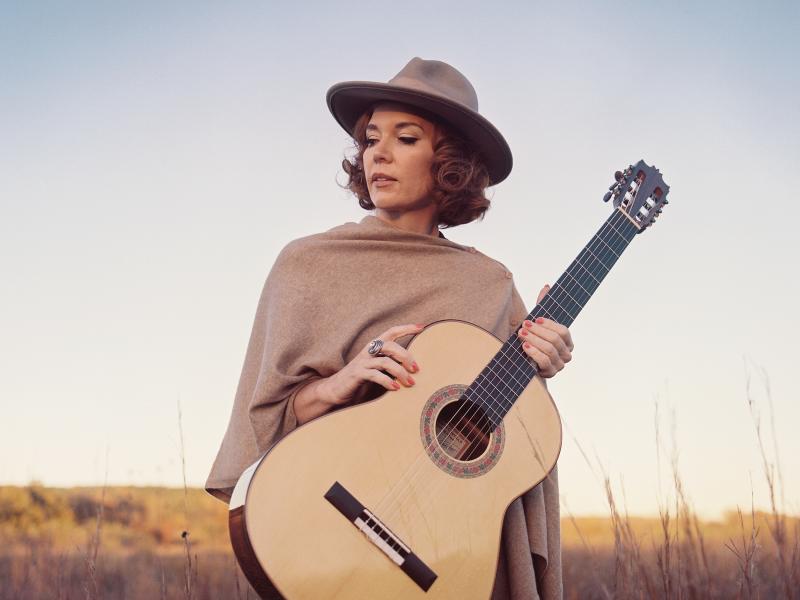 Sue Foley in field of grass looking off to the side in a hat and shall holding an acoustic guitar