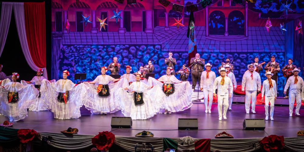 Folklorico dancers dressed in white costumes dance on a stage with mariachi musicians behind them