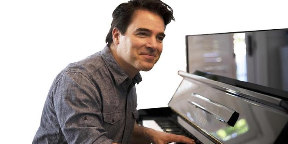 A wearing a grey shirt looks at the camera smiling, while playing the piano