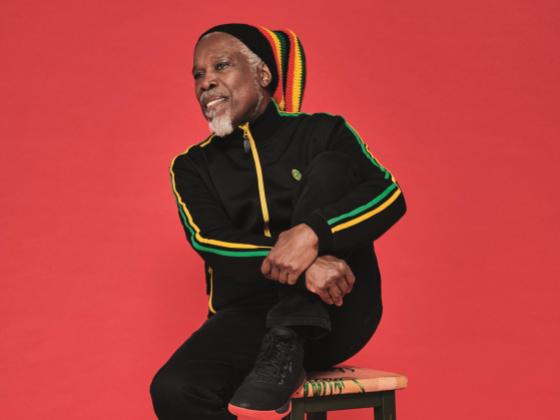 A man with a grey beard sits on a stool, wearing a black track suit and a Rastafarian-colored beanie hat