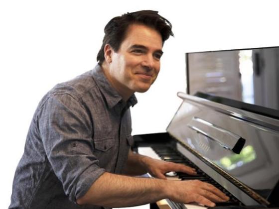 A wearing a grey shirt looks at the camera smiling, while playing the piano