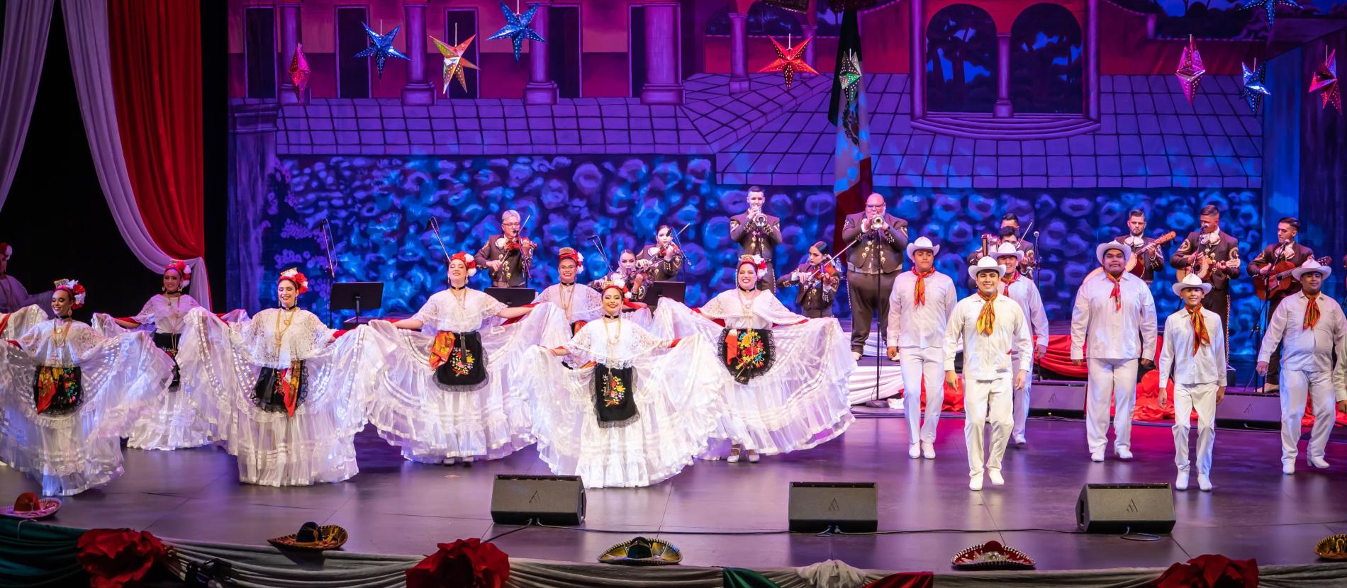Folklorico dancers dressed in white costumes dance on a stage with mariachi musicians behind them