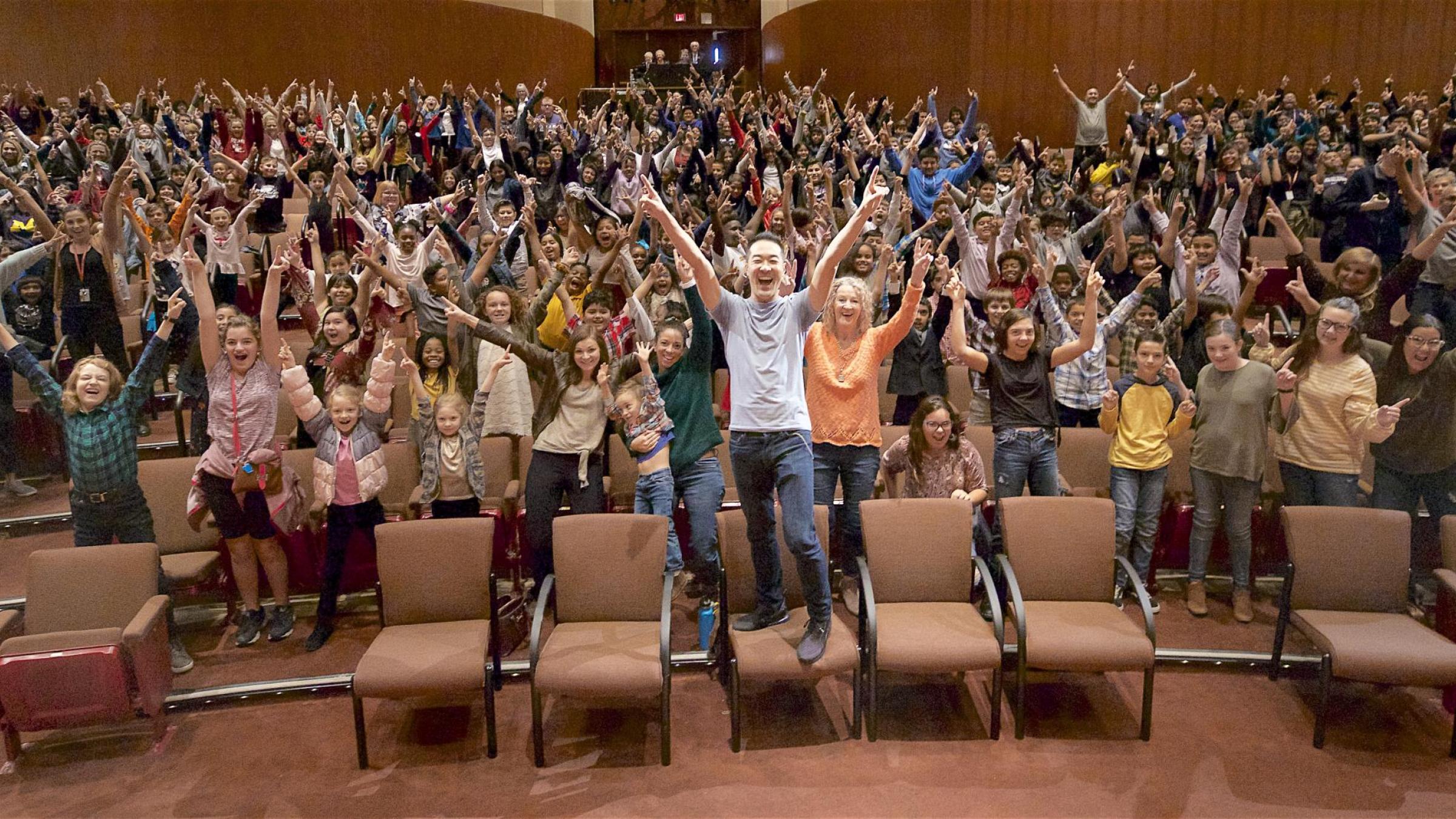 Artist Alpin Hong stands with his hands raised in joy along with a theatre full of children