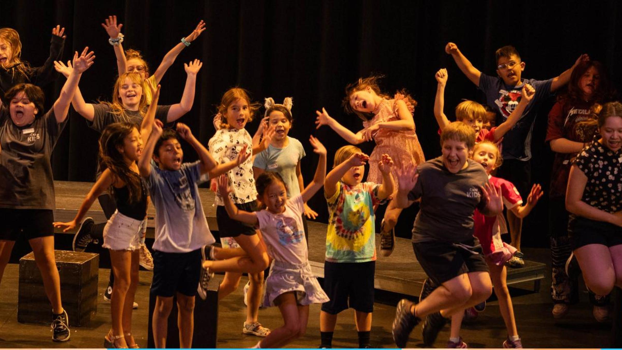 A group of youth on the theatre stage, all in a moment where they are jumping up in the air and making silly faces